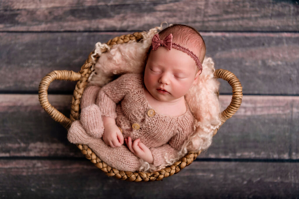 Newborn girl in dusty rose knit outfit and headband sleeping in wicker basket on rustic wood backdrop