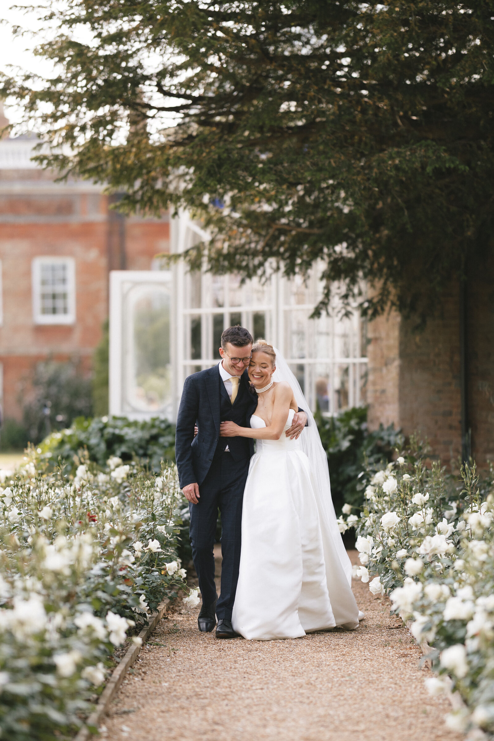 A bride in a white gown and veil walks arm in arm with a groom in a dark suit along a garden path lined with white flowers, both smiling happily under a large tree at their romantic Avington Park Wedding.