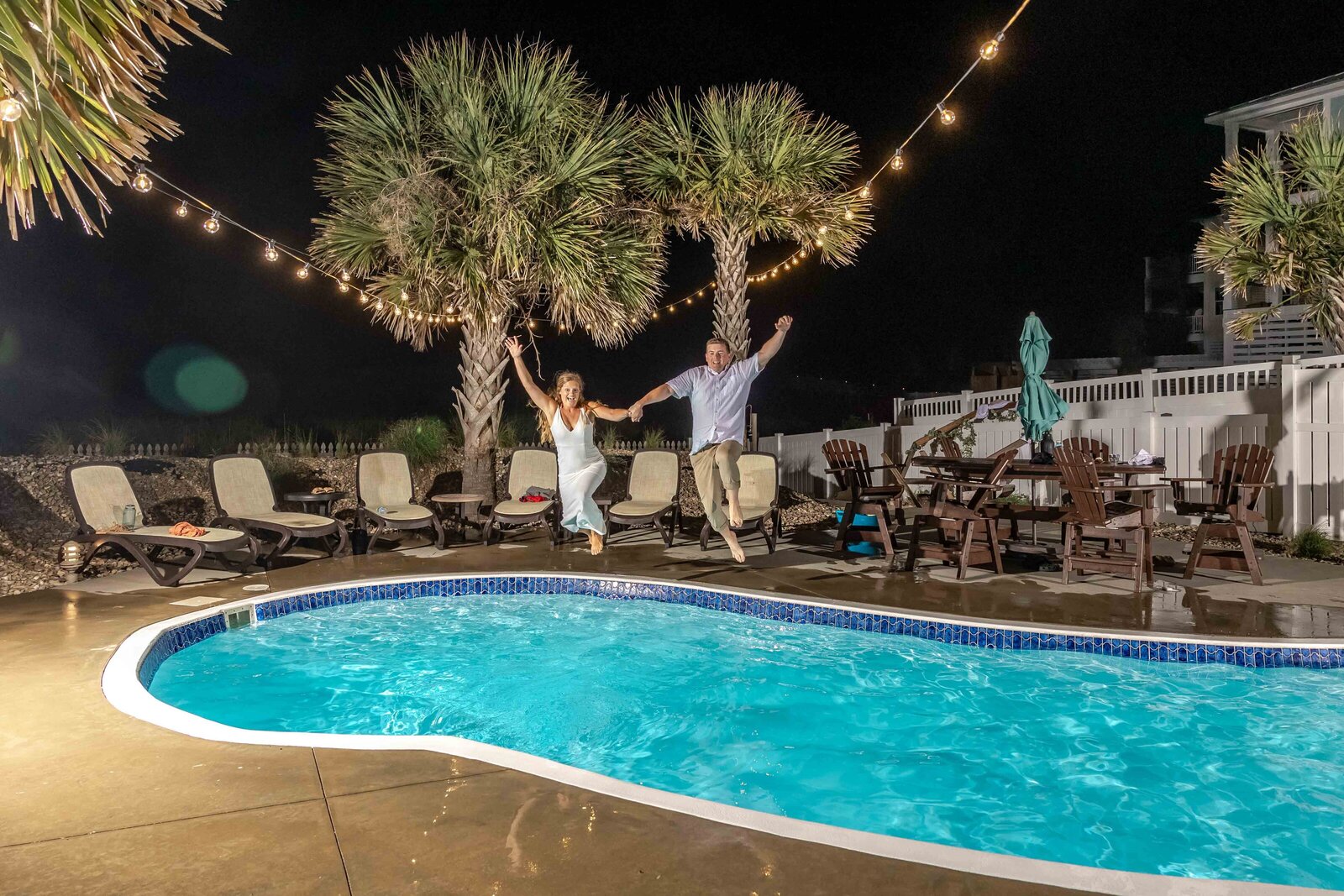 The bride and groom hold hands and jump into a pool under string lights during a fun and playful Topsail Beach, NC wedding, blending coastal charm and spontaneous celebration.