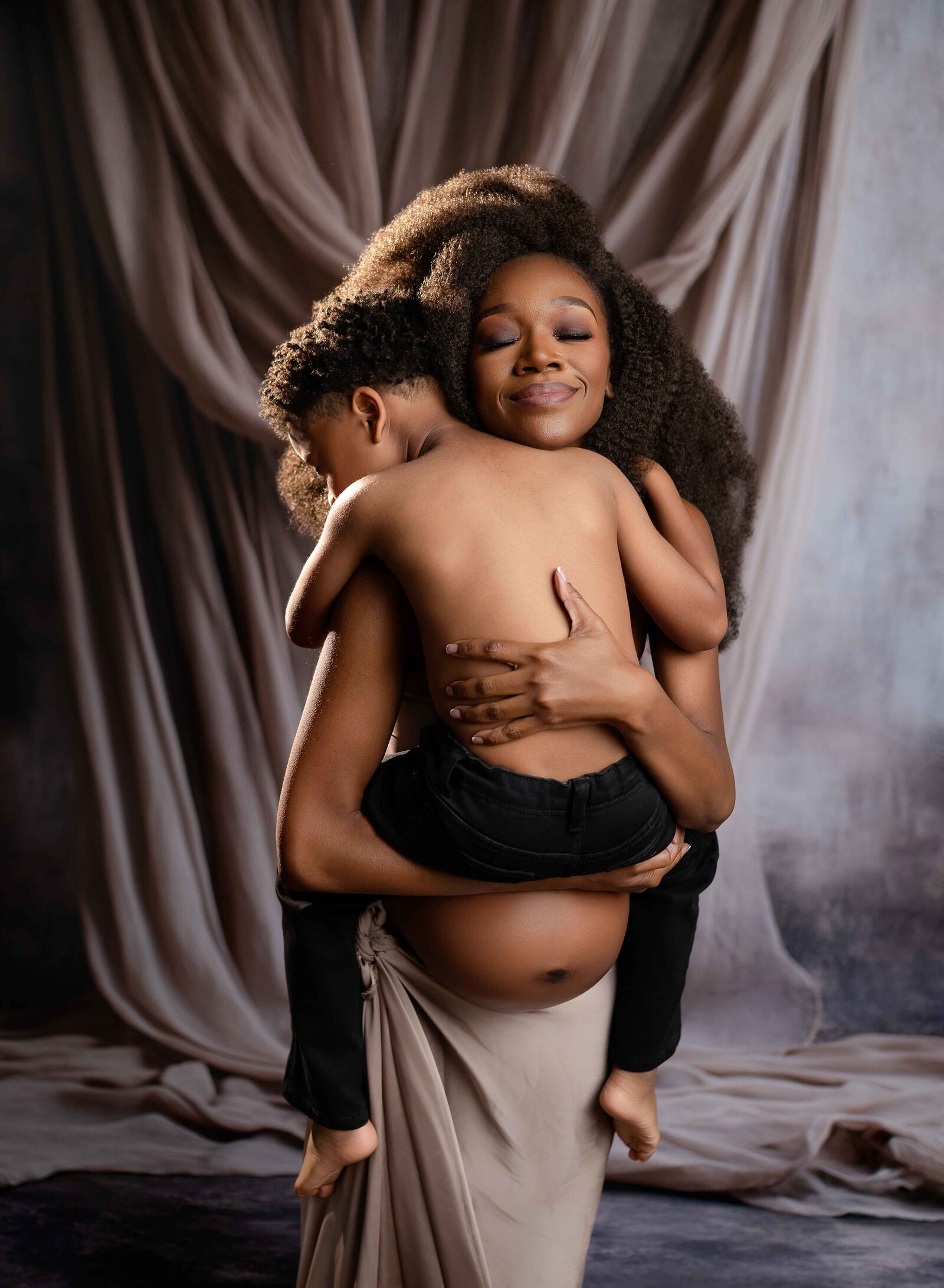 Pregnant women with baby bump out, hugging older toddler son and smiling with eyes closed. Soaking in the final moments of motherhood before 2nd baby is born.