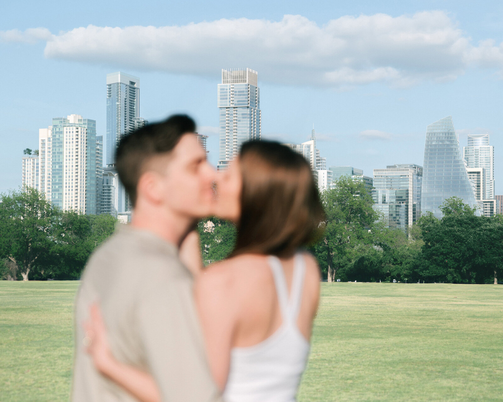 engagement session with austin downtown skyline in the background. man and woman are kissing and out of focus in the foreground, austin skyline is in the focus for this engagement photo