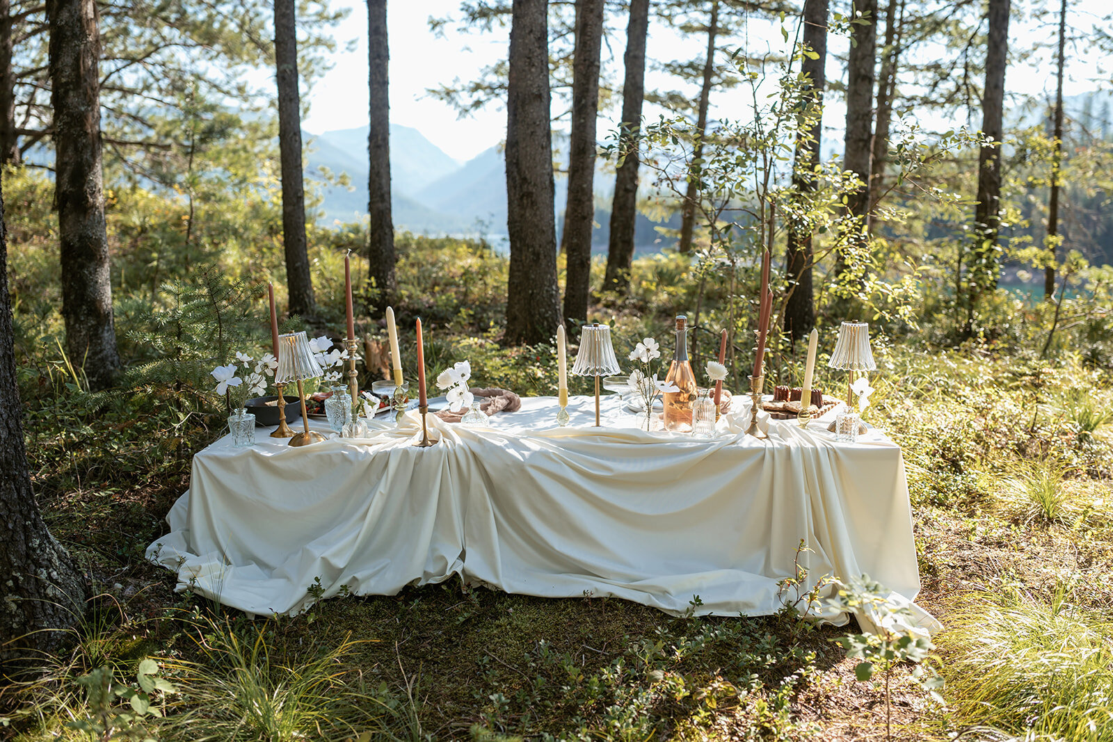 A dreamy elopement dinner table in the woods, styled with soft draped linens, candlesticks, and delicate florals under golden afternoon light.
