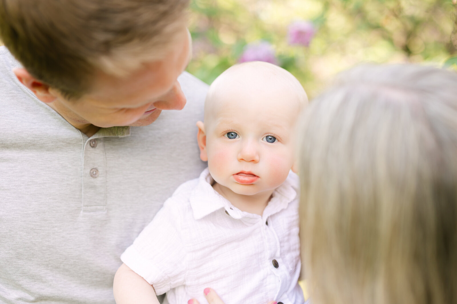 a baby boy is held between his mother and father, gazing at the camera with his bright blue eyes while wearing a white button up shirt
