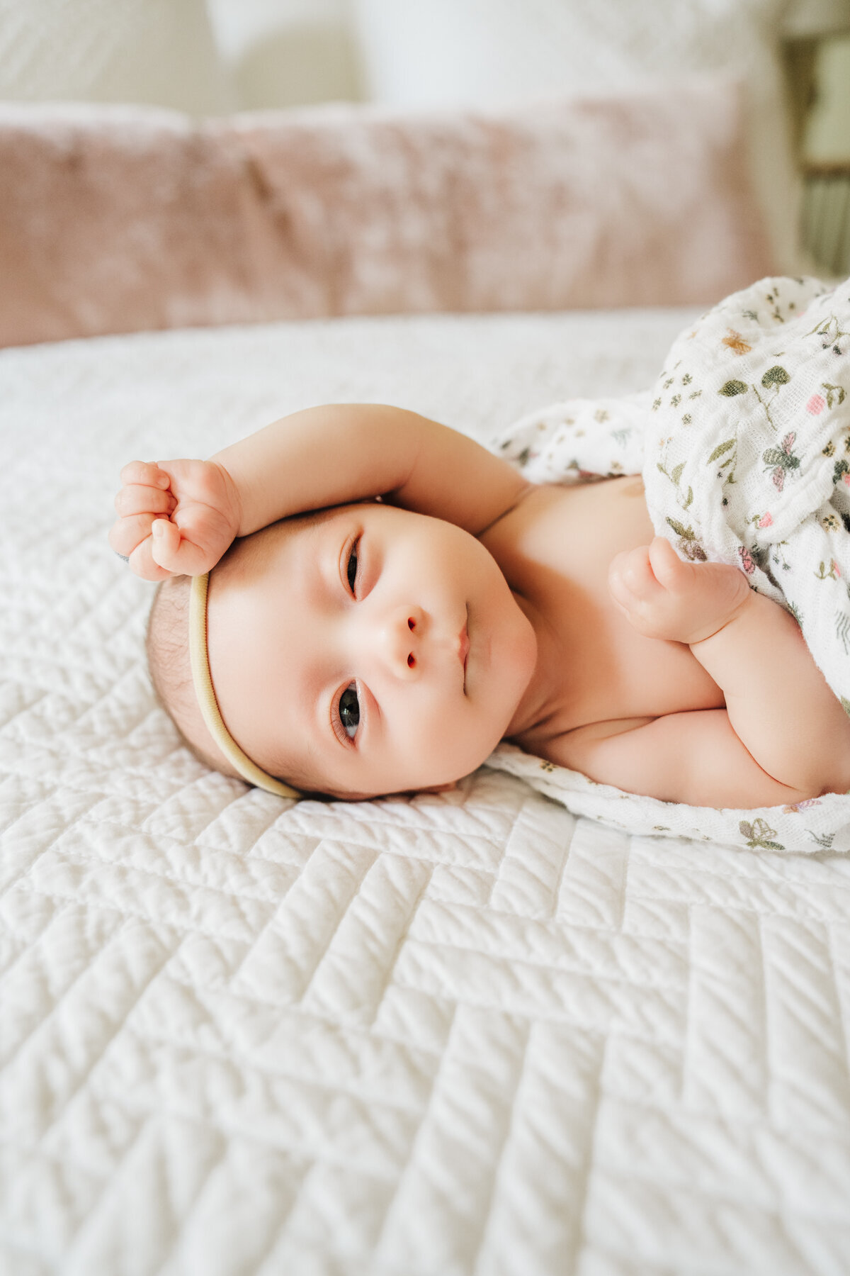 newborn girl in a floral wrap winks at camera while laying on white bedspread