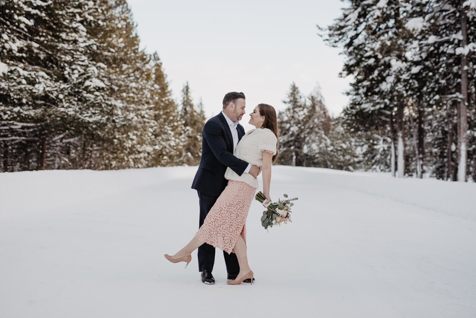 grand teton wedding with groom holding his bride who is wearing a pink wedding dress as they smile into each others eyes as the bride holds her floral bouquet to her side with the trees in the distance down the snowy road