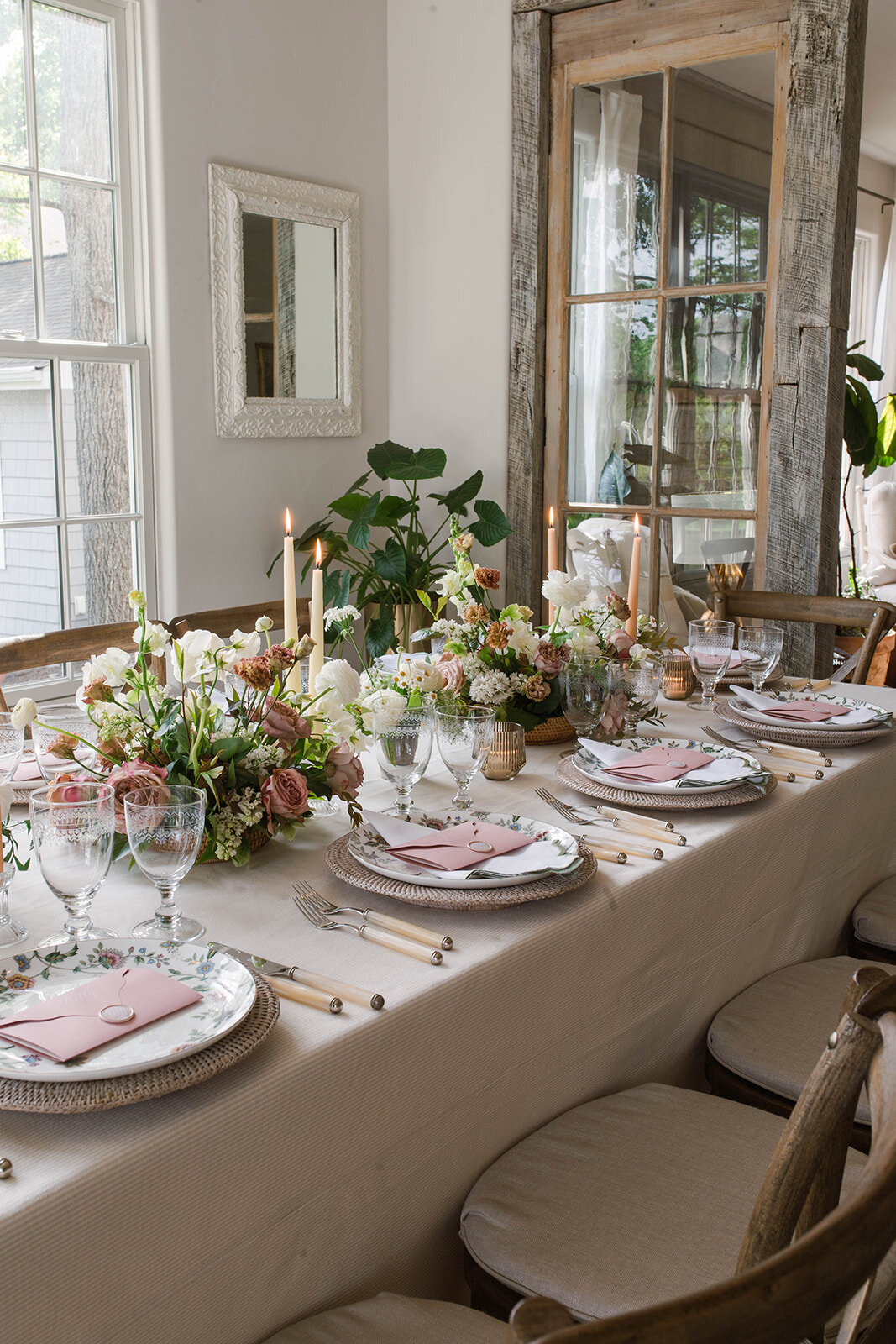 A long table with floral arrangements including white ranunculus, white snapdragons, brown lisianthus, mauve garden roses, and greenery with cream and sand-stone colored taper candles.