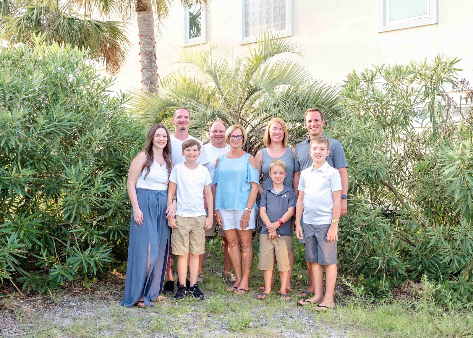 A family poses in front of lush greenery and palm trees, dressed in casual summer attire. The relaxed setting highlights their close bond. Perfect for professional family photography in NC, showcasing natural and intimate family moments.
