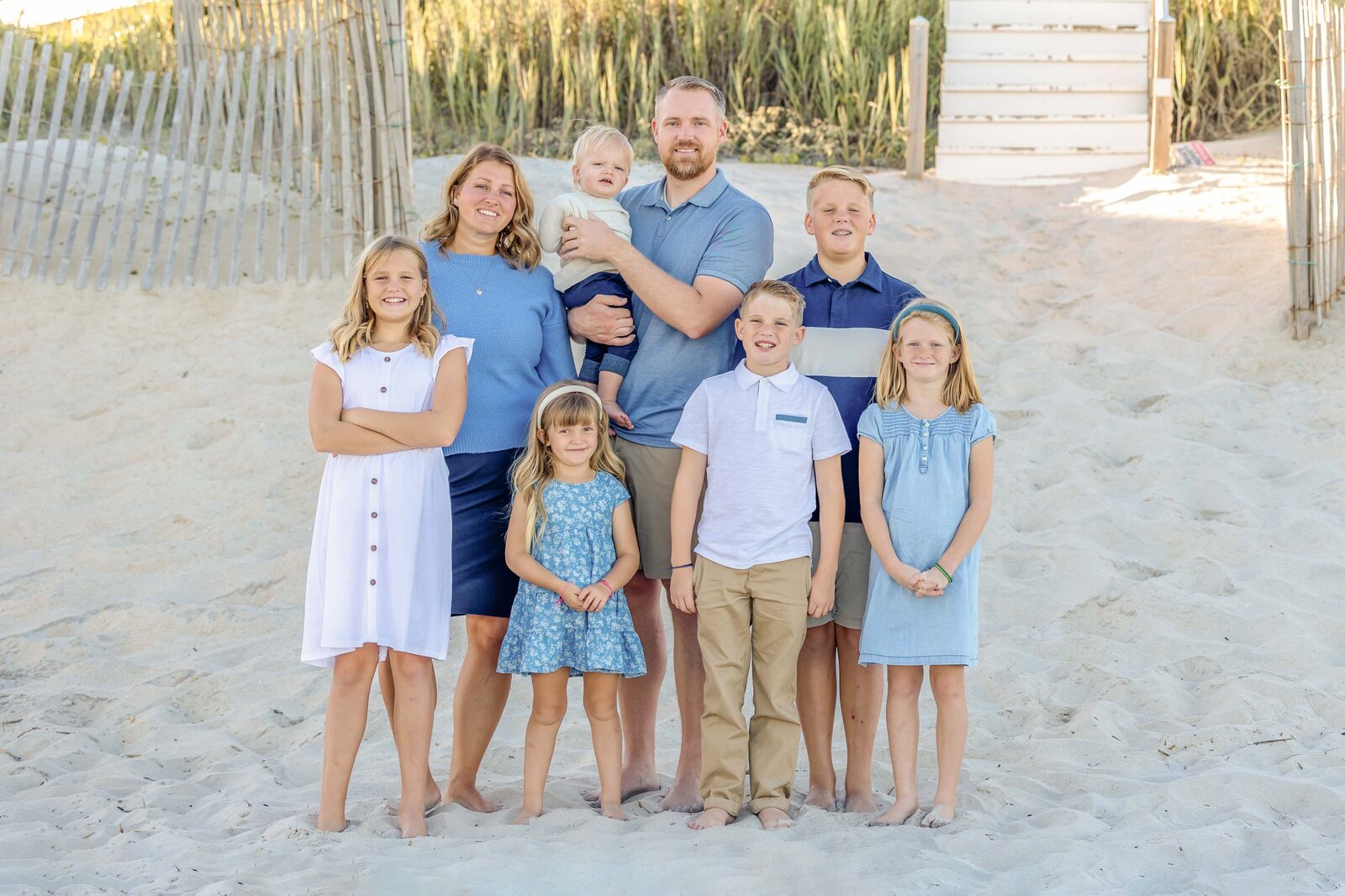 A family of seven poses on a sandy path leading to the beach, dressed in coordinating blue and white outfits. The wooden fencing and grass create a classic coastal setting. Perfect for artistic family photography in Wilmington, NC, capturing family unity.