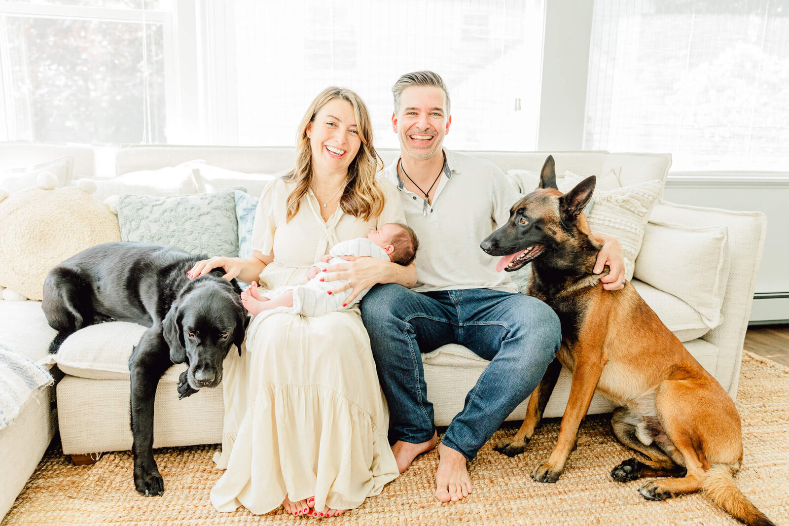 Mom holds newborn and smiles with dad while two dogs sit nearby