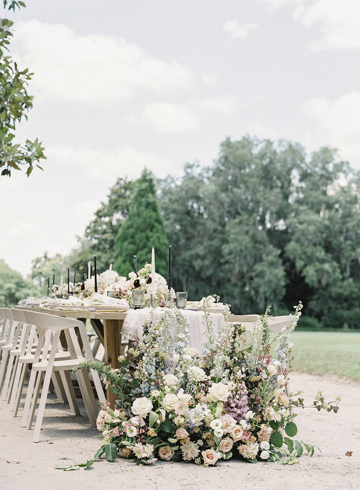 Outdoor reception table with floral installation from ground to top of the table. Flowers are in deep purples, pinks, greens, ivory and taupe. Kings table with 16 guest spots, cream colored chairs, hand dyed linens running down the center of the table. Flowers continue down the middle of the table with black and white candles. Photographed at Middleton Place by wedding photographers in Charleston Amy Mulder Photography.