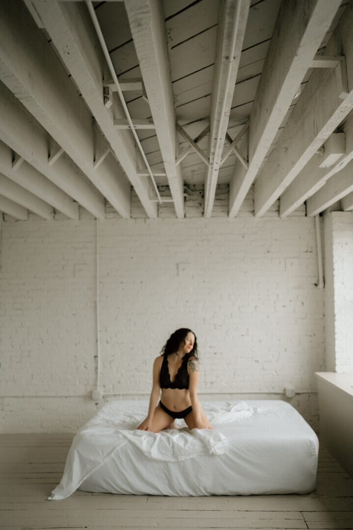 woman kneeling on a white bed wearing a black lingerie set from Linger Boutique in Olympia WA looking out the window