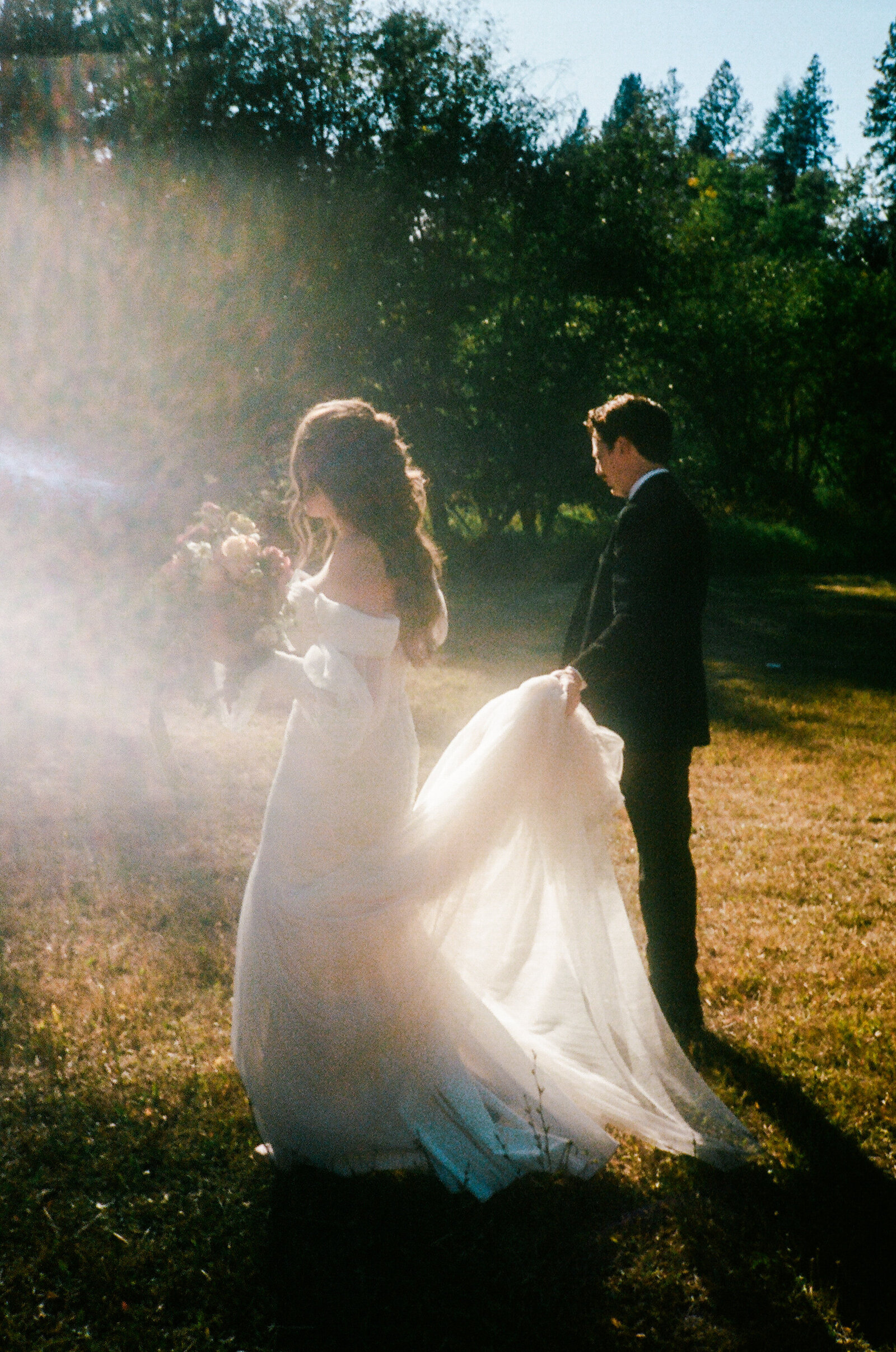 Groom carrying brides dress as they walk along the grass at California wedding