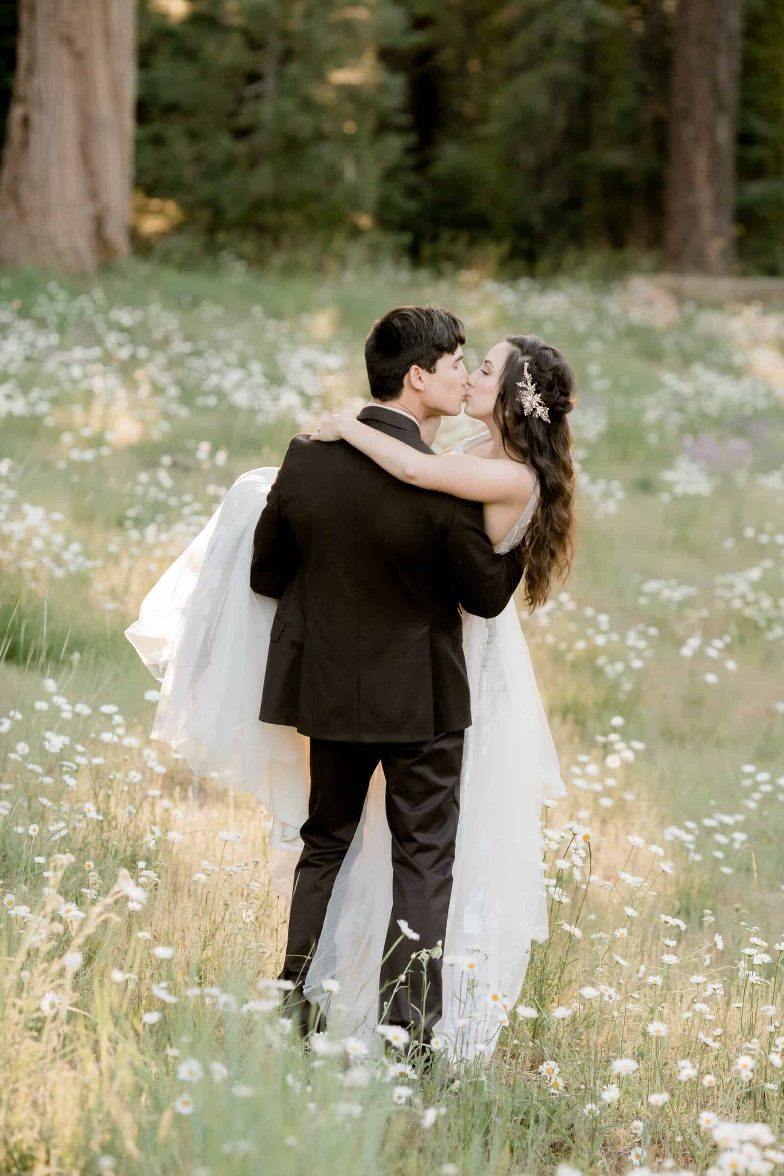 A groom carries his bride in a beautiful meadow of daisies on their mountain wedding day in California.