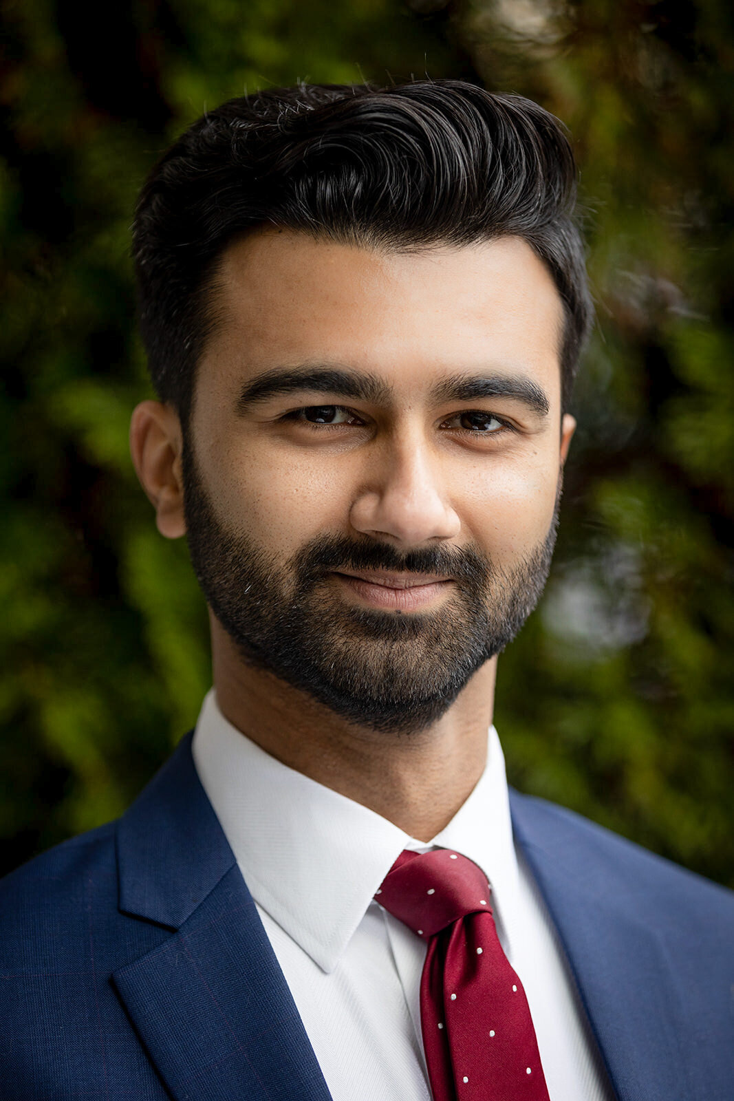 Professional headshot of a man in a navy suit, white shirt, and red tie, posing confidently outdoors with a green, blurred background.