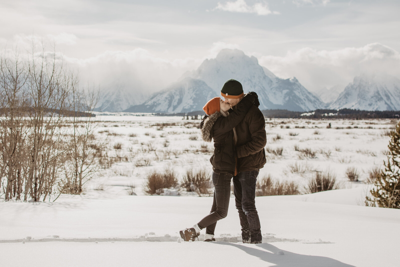 Jackson hole photographers photographs winter engagement in the Grand Tetons with woman looks up to her fiance and kissing him as he leansdown to hold her waist with the mountains in the background