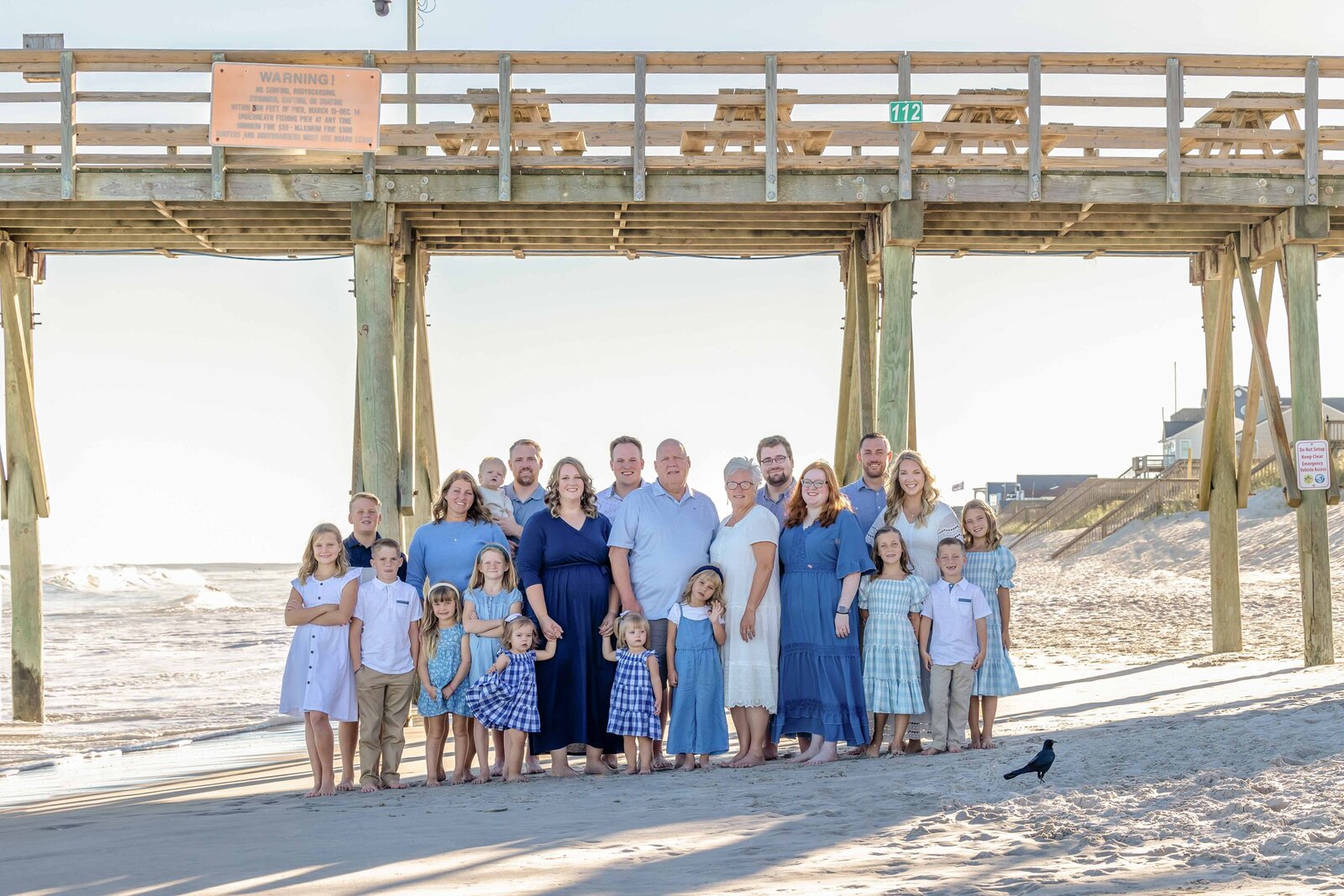 An extended family stands under a pier, dressed in coordinated blue and white outfits. The ocean waves and wooden structure create a picturesque backdrop, ideal for destination family photographer NC, showcasing unity and seaside charm.