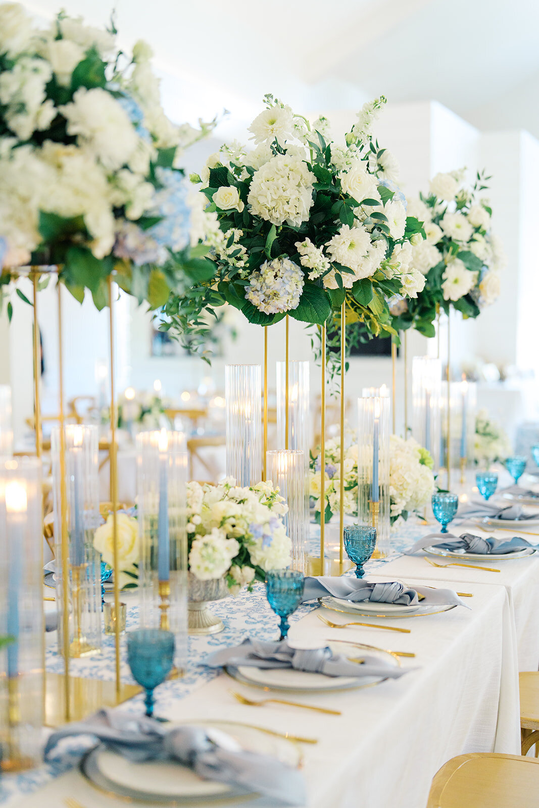 Blue and green wedding tablescape with floral arrangements on stands, blue taper candles and blue goblets.