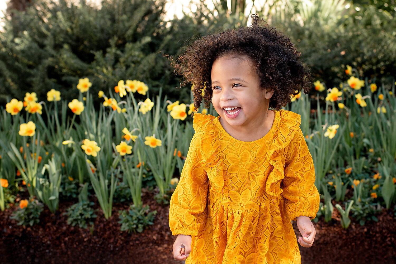 A young girl with curly hair wearing a yellow dress and smiling and laughing standing in front of yellow flowers.