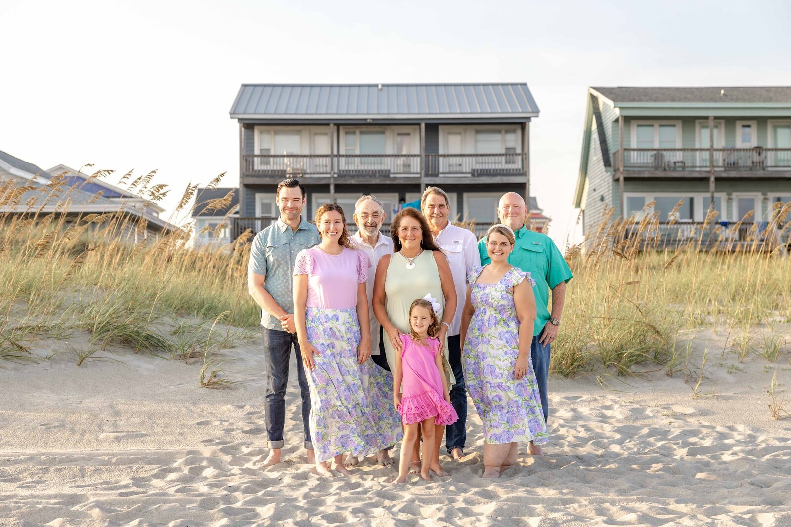 A family stands on the sandy beach in front of coastal homes, dressed in pastel and floral outfits. The sea oats and bright sky create a charming coastal scene, ideal for artistic family photography in Wilmington, NC, capturing relaxed beachside moments.