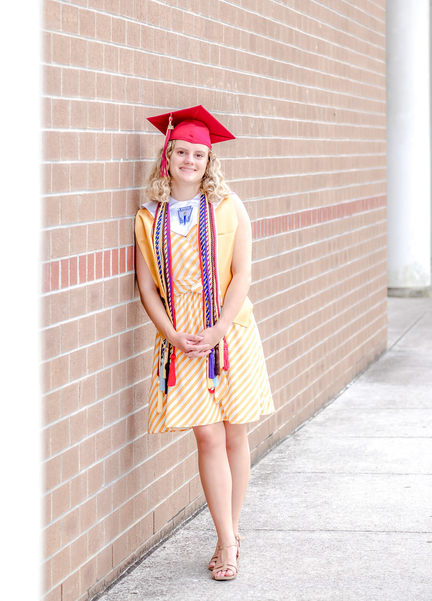 An Ashley High School senior in a red cap and gown, adorned with honor cords, leaning against a brick wall in Wilmington, North Carolina. This portrait captures the pride of graduation, ideal for those seeking professional senior photography.