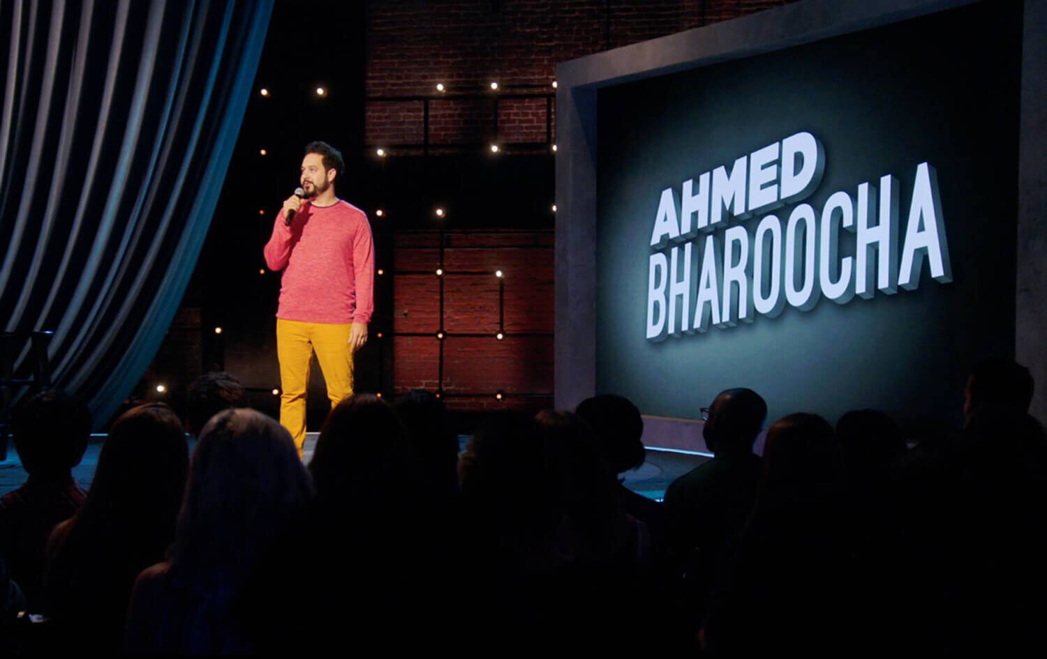 Photo of comedian, Ahmed Bharoocha, on stage and holding a microphone with his name off to the right