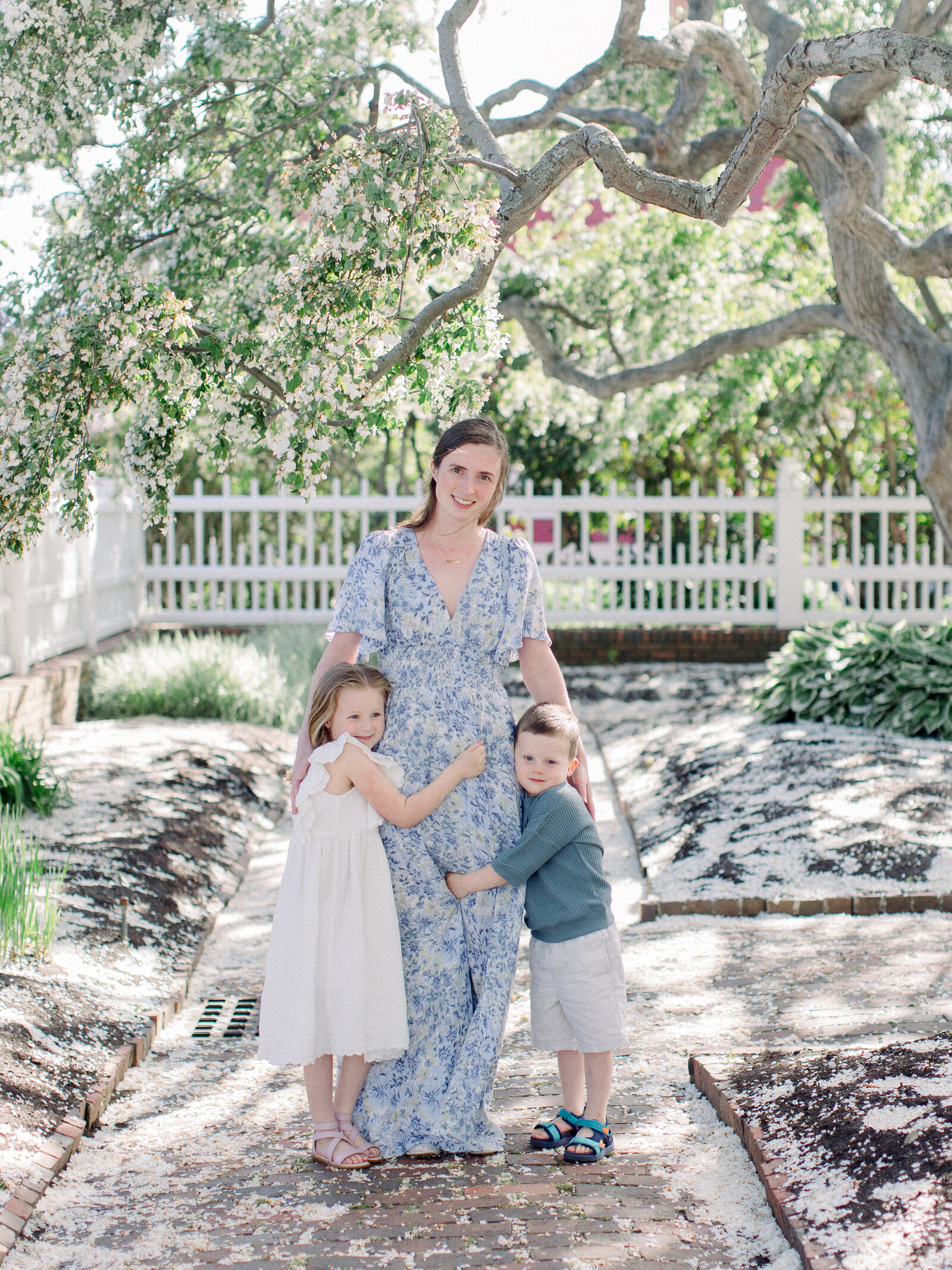 Mom in a blue and white floral gown hugging her two children by her side in a spring garden with flowering cherry trees by NH newborn photographer Fieldstone Studio.