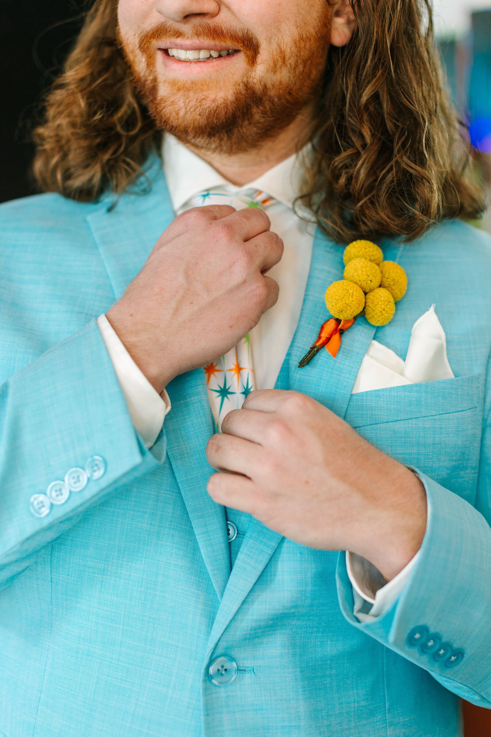 A groom in a light blue suit adjusting his mid-century starburst patterned tie with a boutonniere made of 5 yellow billy buttons