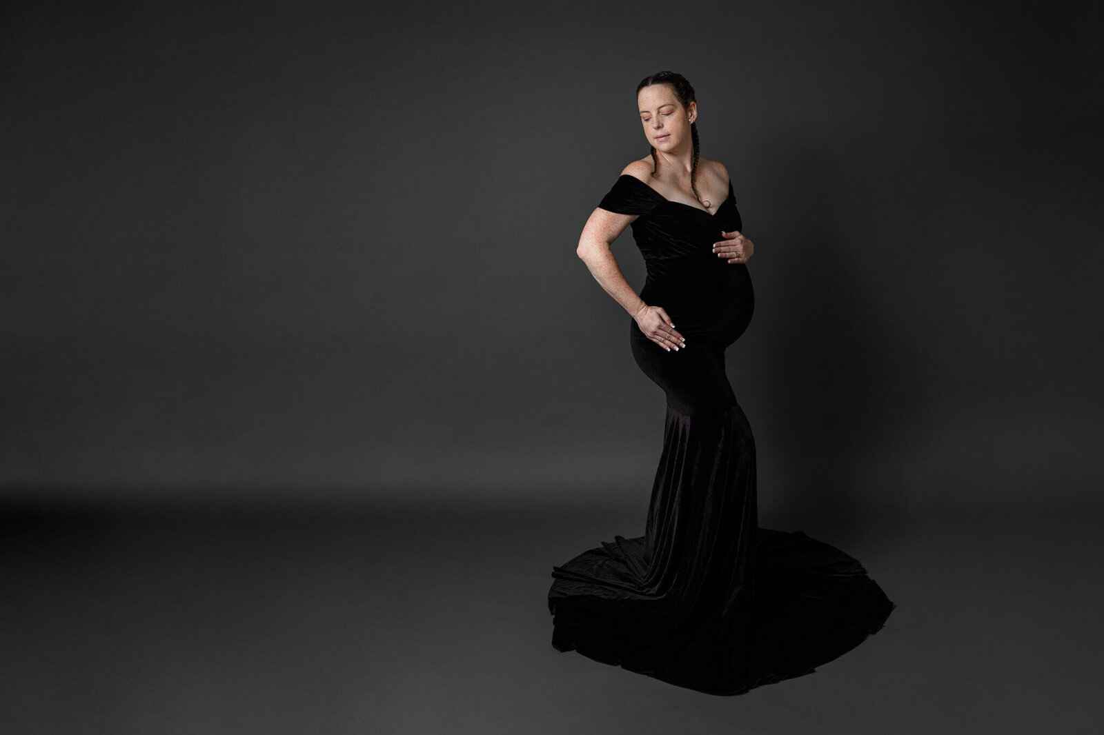 A pregnant woman in a black dress posing on a gray backdrop.