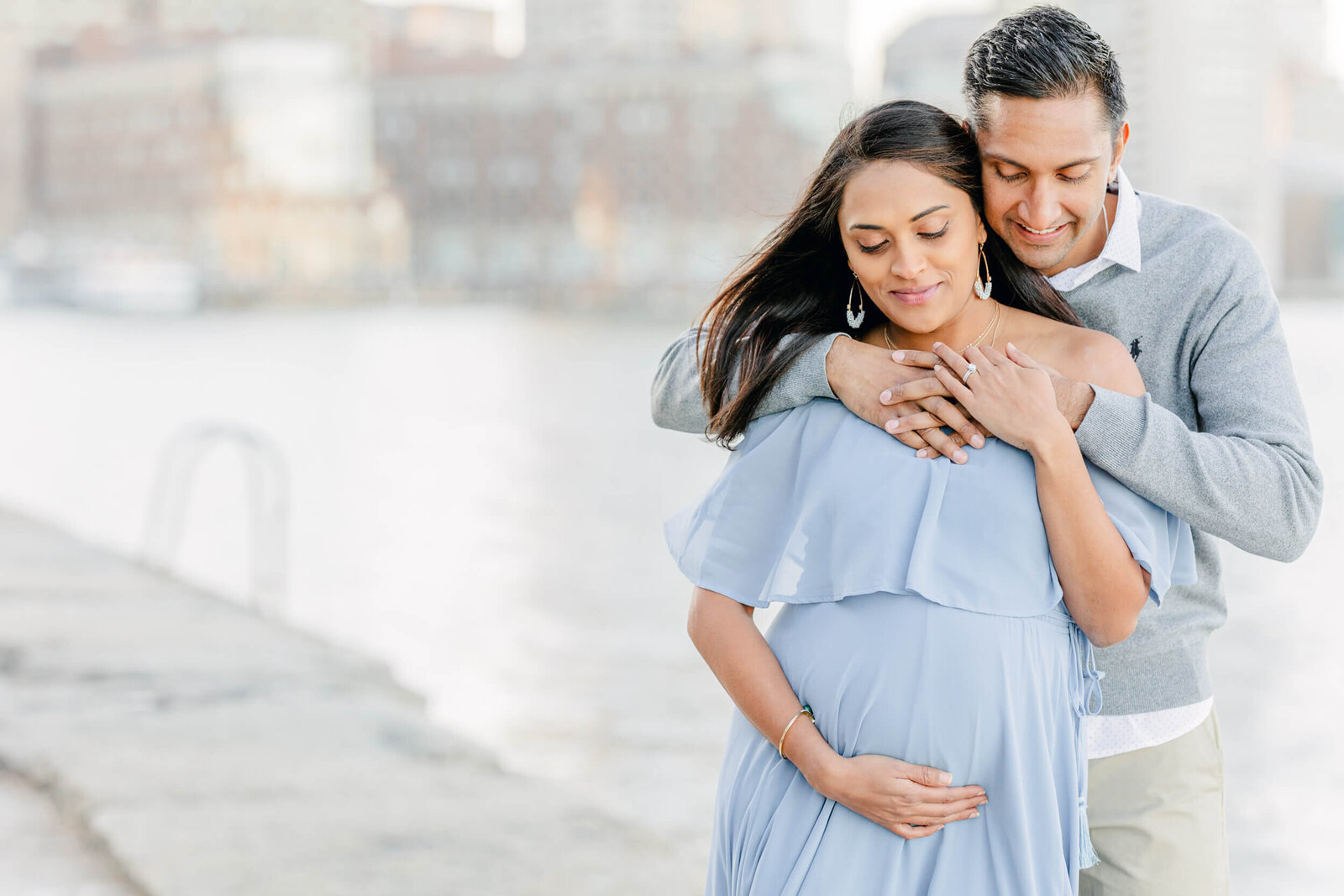 Pregnant woman in a flowing blue dress smiles while her husband hugs her from behind