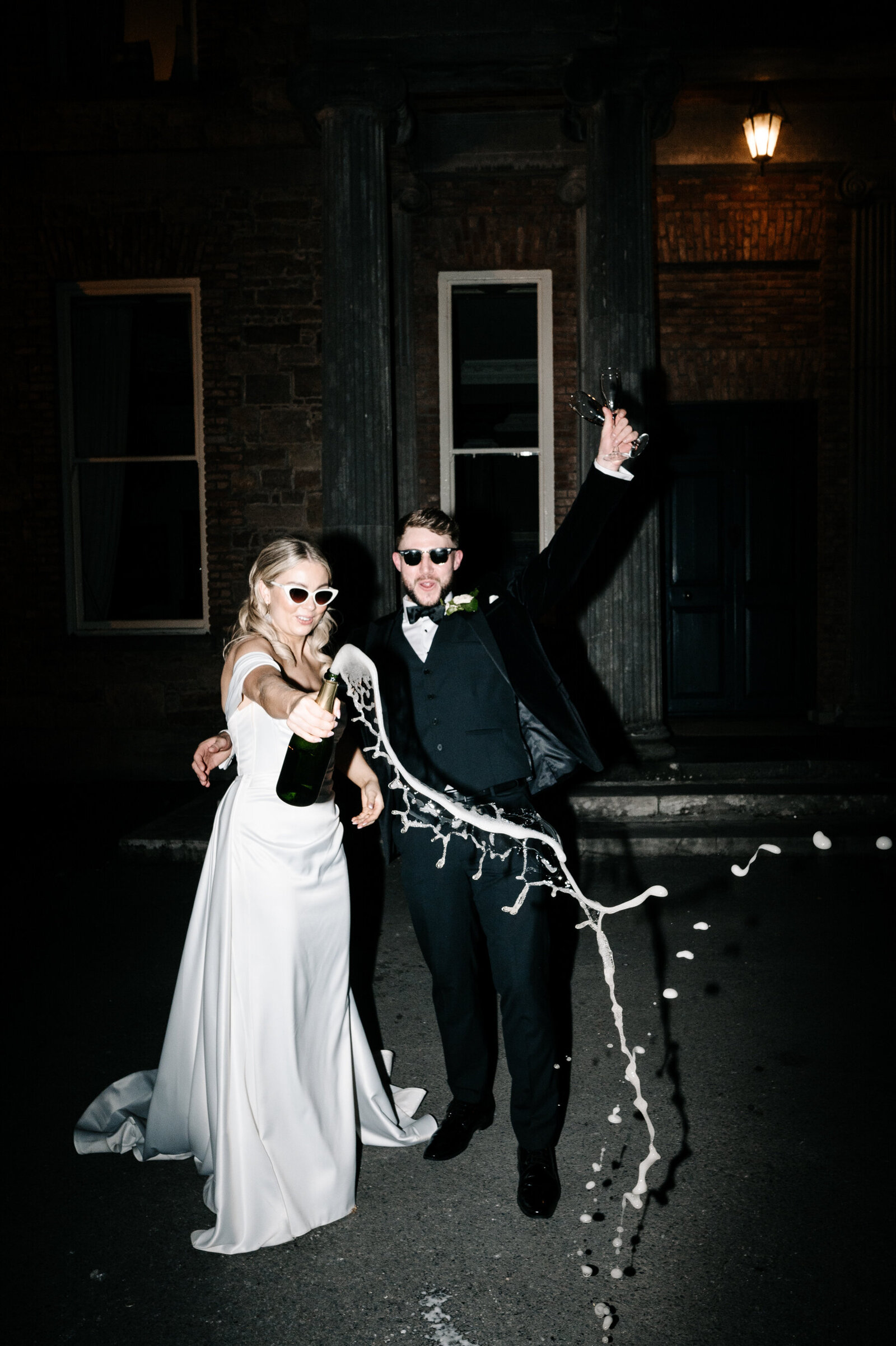 Bride pops champagne bottle while groom celebrates in the background