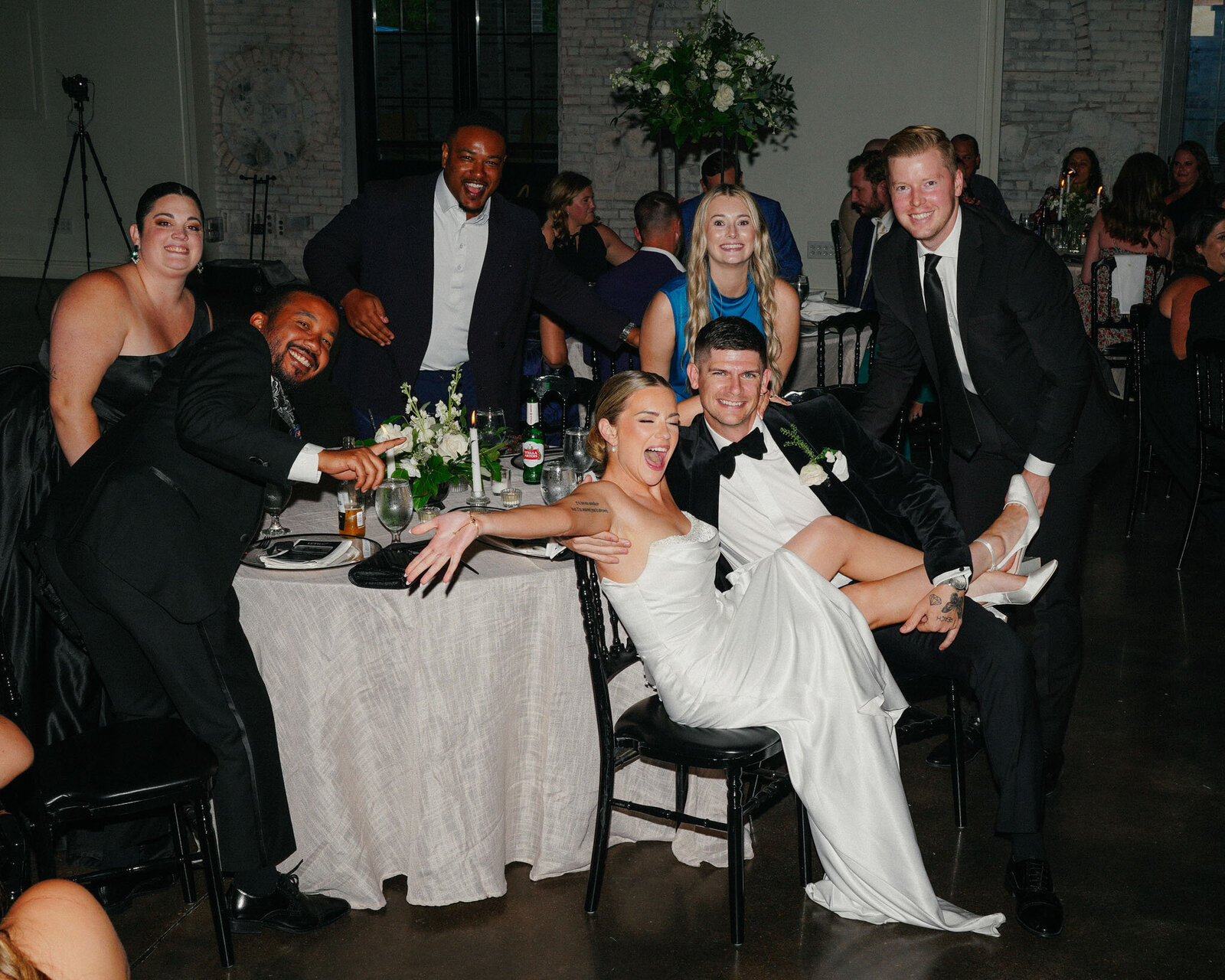 A bride and groom happily share a playful moment together as their guests gather around them, laughing, cheering, and striking fun poses during the celebration.