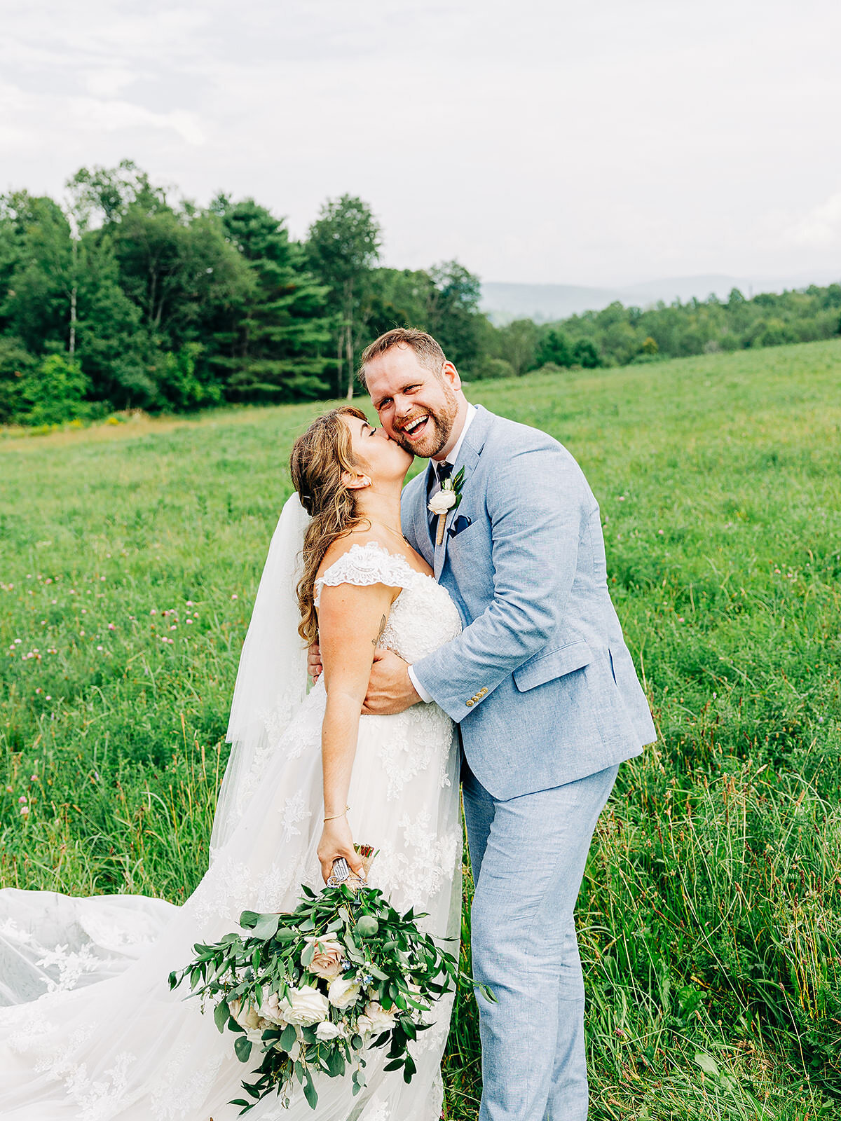 A bride and groom smile joyfully while embracing in a lush green field, captured beautifully by an Eau Claire Wedding Photographer. The bride holds a bouquet of white flowers with trees and distant hills in the background.