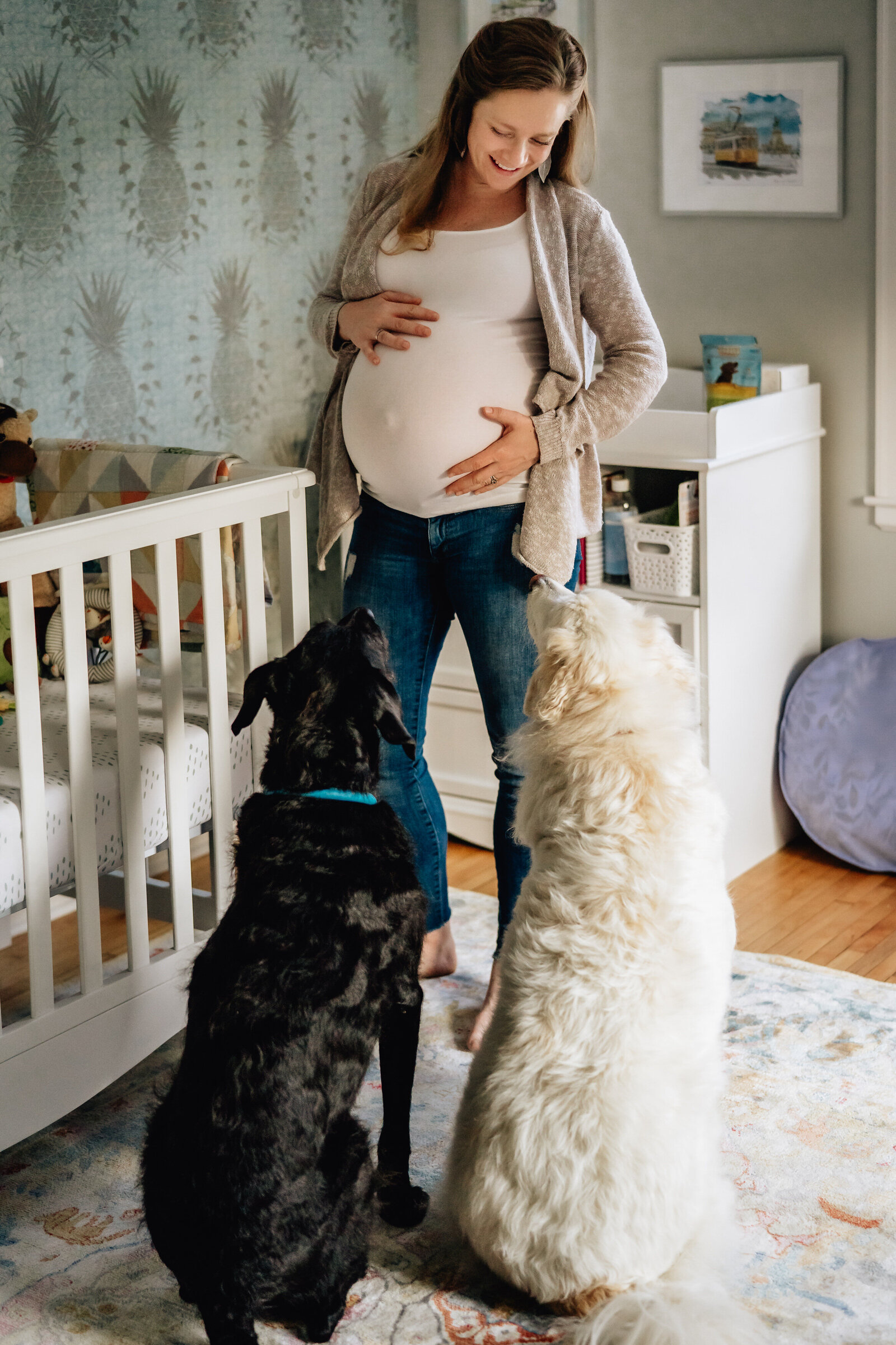 A pregnant woman stands in a nursery, gently holding her belly while looking down at two dogs sitting in front of her. The room is softly lit and decorated in a cozy, warm style.