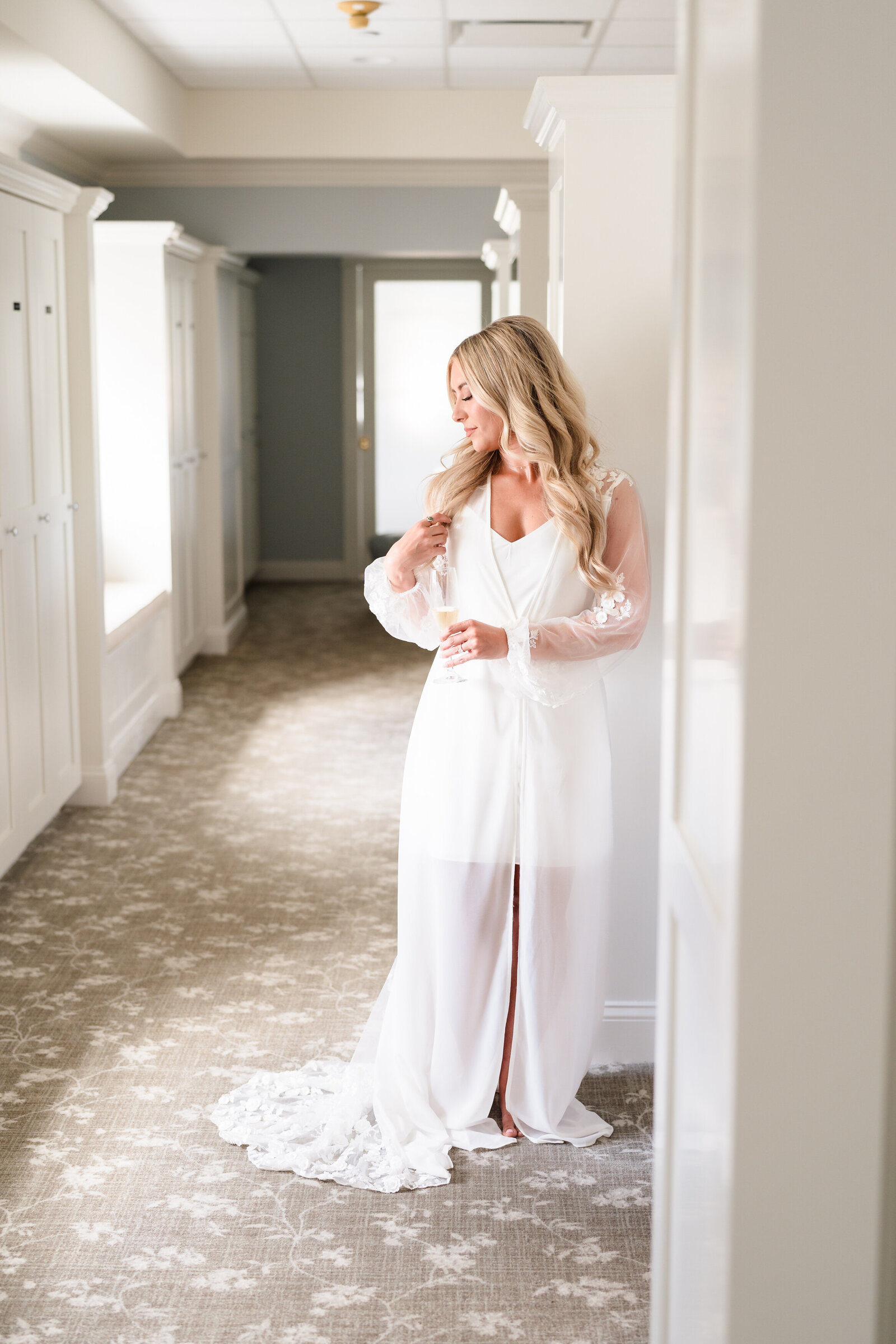 Bride holds a glass of champagne and strokes her hair as she stands in a window-light hallway wearing her getting ready robe