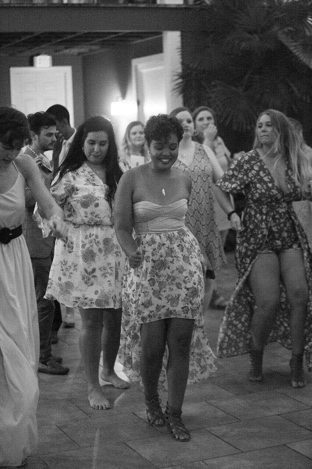 A group of people dance together outdoors, most wearing floral dresses. The image is in black and white, and everyone appears to be enjoying themselves on the dance floor.