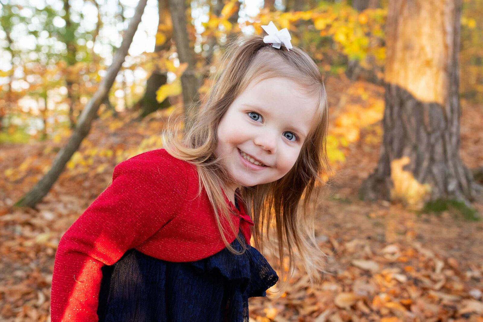 A little girl in a black dress with a red cardigan standing among the fall foliage.