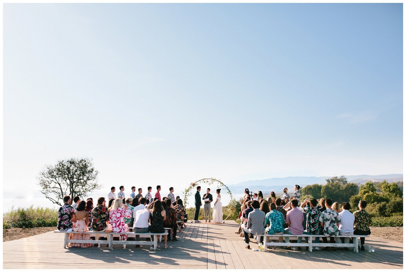Full wedding at Dos Pueblos Orchid Farm.  The bride and groom are sttanding under a circle arch with their bridal party on the sides. There are a lot of guests looking. The view overlooks some cliffs and ocean