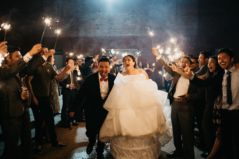 A joyful bride and groom walk through a cheering crowd of guests holding sparklers at night. The bride wears a voluminous white gown, and everyone is smiling and celebrating their wedding.