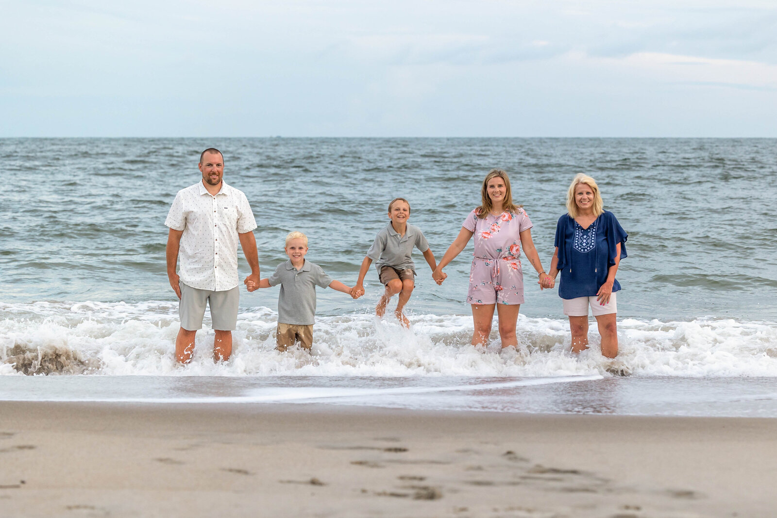 A family of five holds hands and plays in the surf at Carolina Beach, dressed in casual summer attire. The playful interaction with the waves highlights joyful family connections. Perfect for affordable family photography in Wilmington, NC.