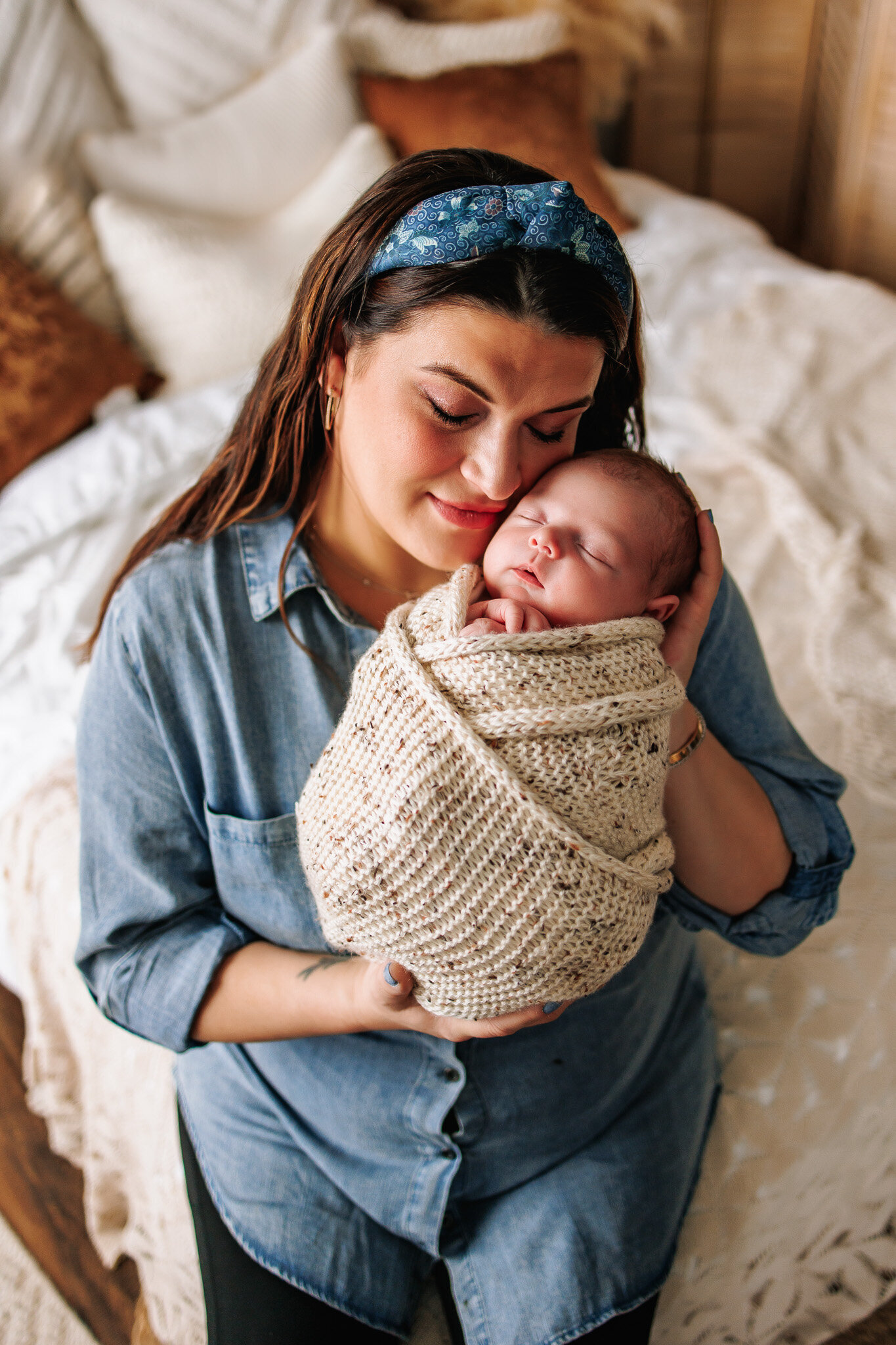 Mother wearing a blue top snuggles cheek to cheek with her newborn son wrapped in a cream knit wrap
