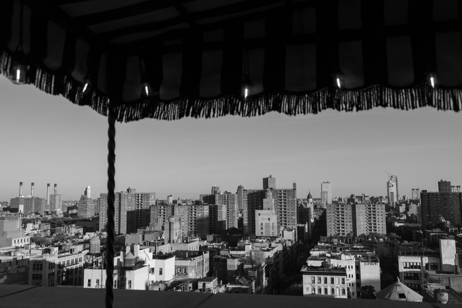 Black & white horizontal photo of NYC skyline with fringe hotel balcony awning and twinkle lights silhouetting the foreground