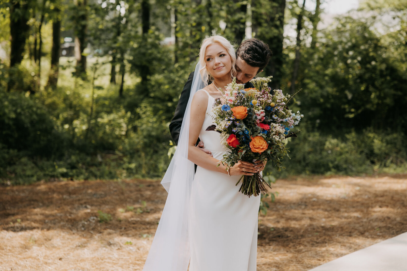 Groom gently kisses his bride's neck while she smiles, standing in front of a wooded area at Kincaid Manor.