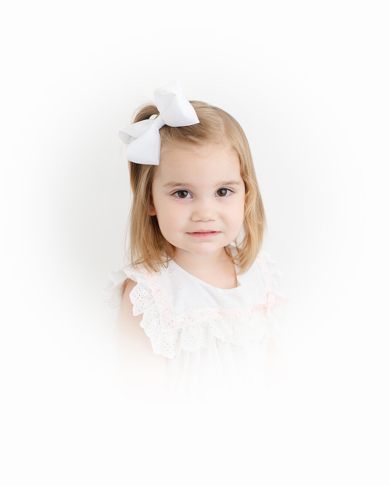 In this portrait, a young child with light brown hair wears a white dress and a large bow, gazing directly at the camera against a soft white background.