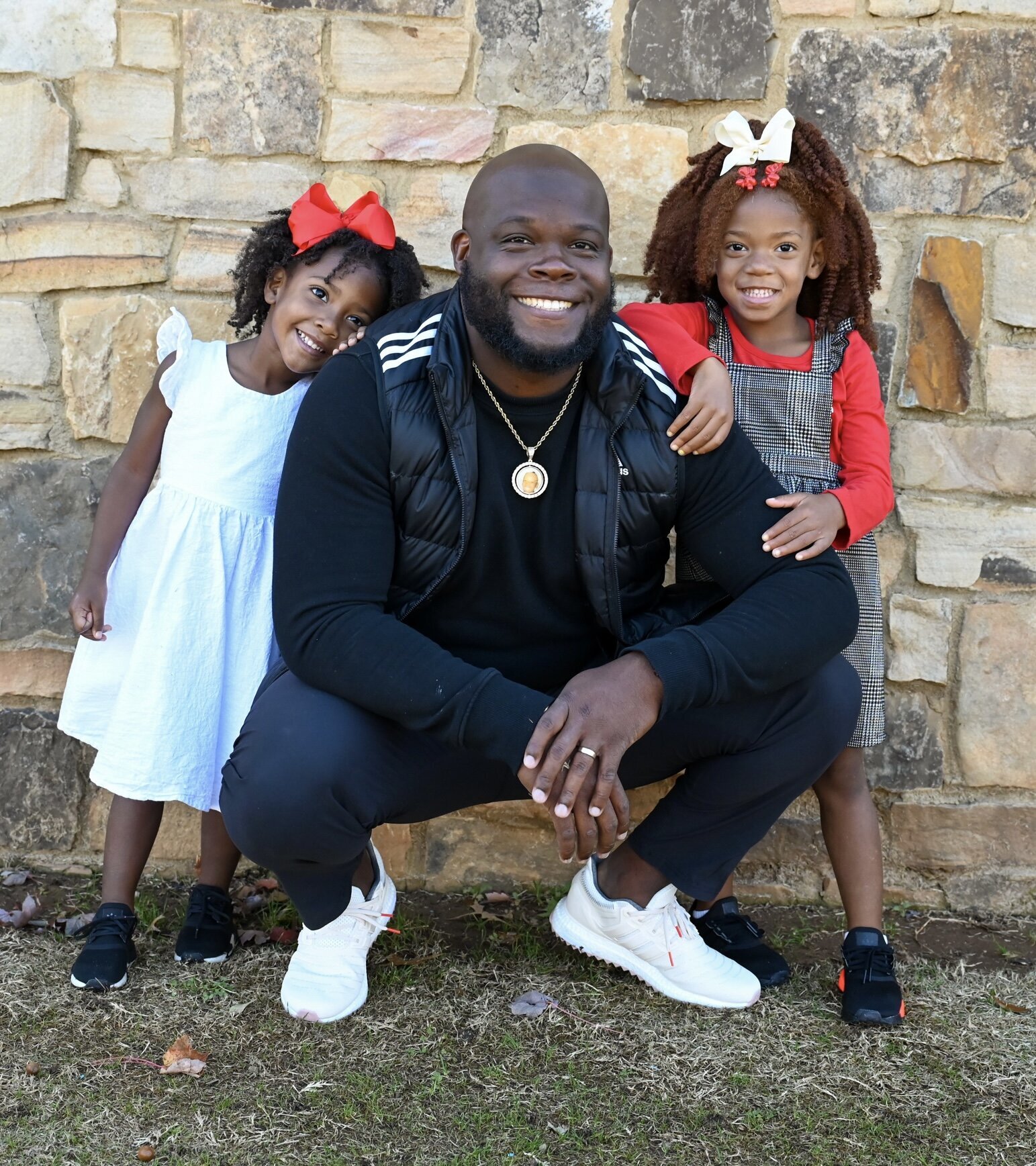 a father posing with his two young daughers. all are smiling at the camera posed in front of a brick wall. photographed by Millz Photography in Greenville, SC