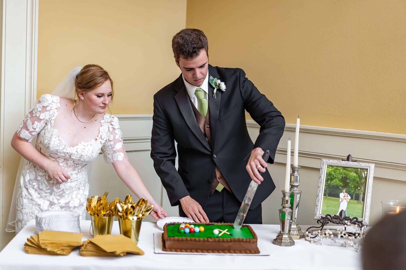 A bride and groom cut a rectangular cake decorated like a billiards table. The table is set with gold utensils, plates, a framed photo of the couple, and two tall candles. Both are dressed in formal wedding attire.