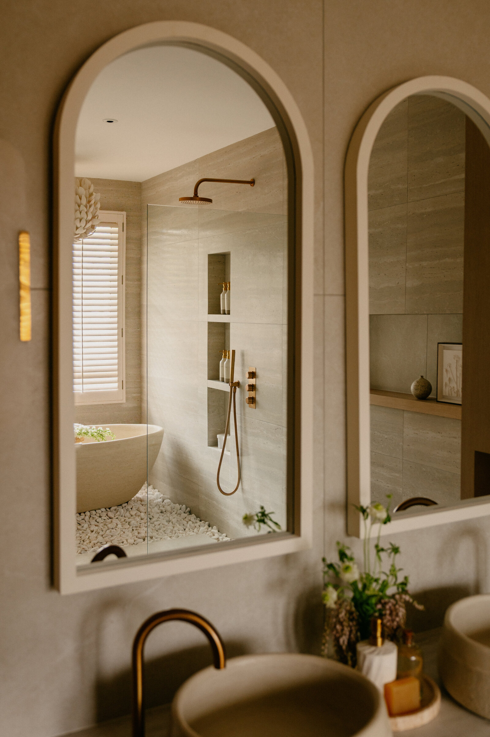 View through arched mirror reflecting stone bathtub, rainfall shower, and pebble flooring beneath shuttered windows.