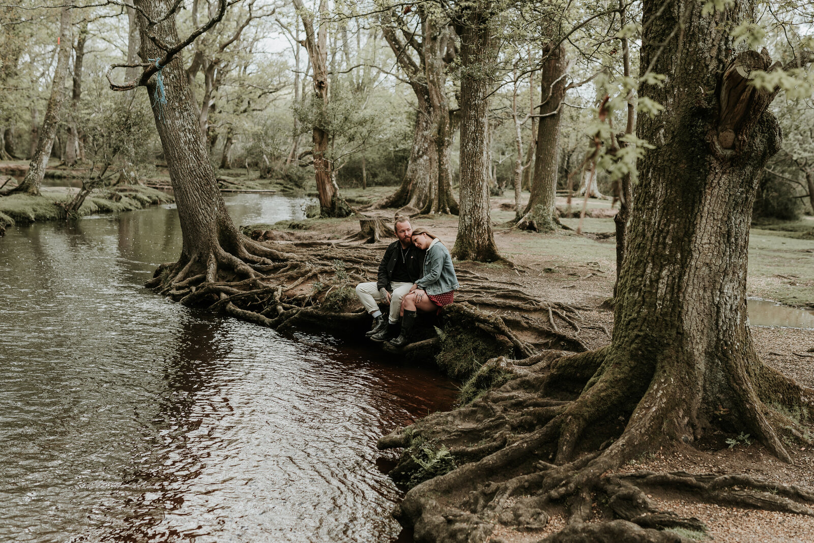 Couple sit along river bank on twisted tree roots in The New Forest