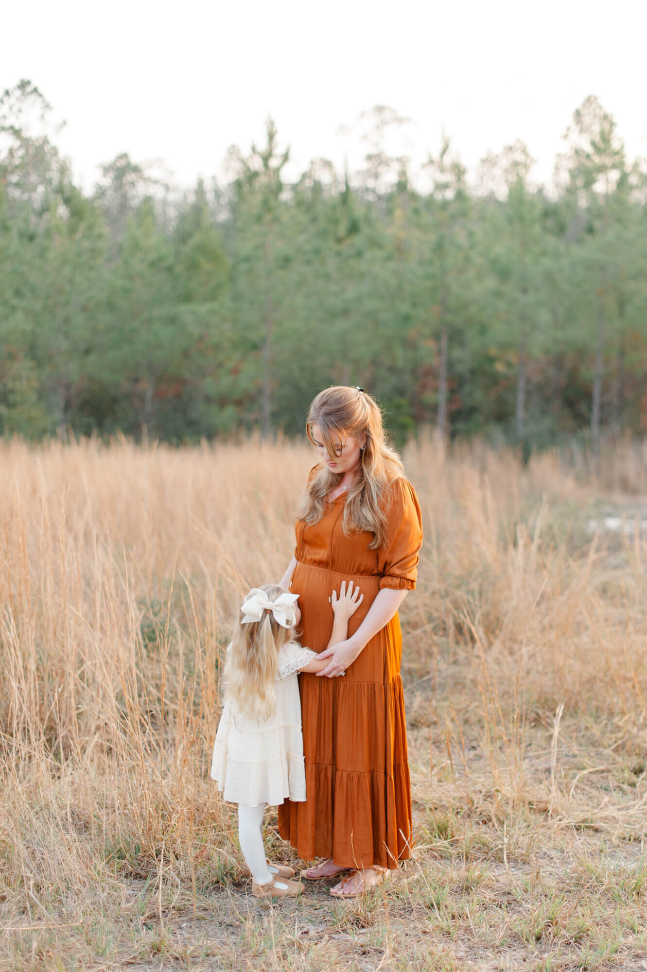 Young daughter stands in a tall grass field with mother kissing her pregnant belly