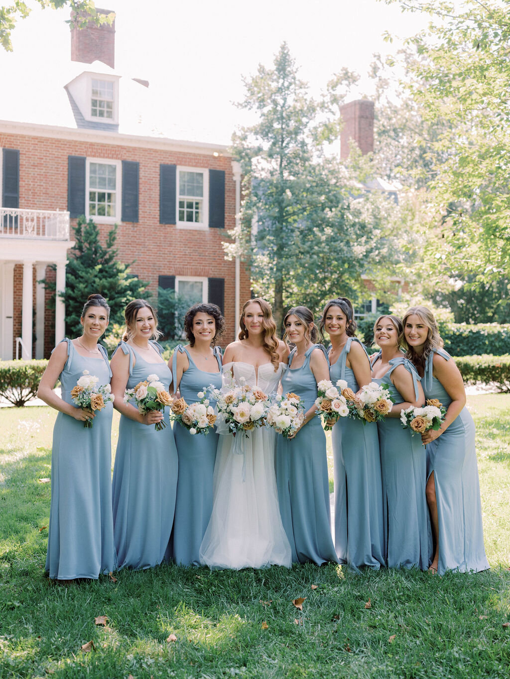 Bridal party holding bouquets in front of the Brittland Estate mansion wearing light blue silk dresses.