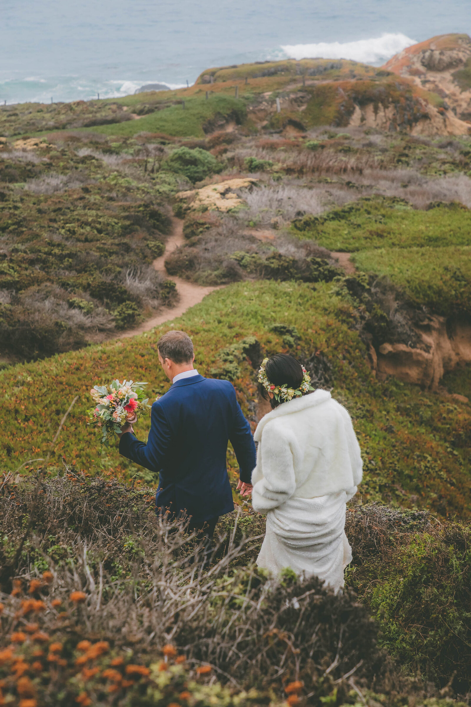 A groom holds the bride's flowers and she wears a flower crown as they hike down a trail in Big Sur.